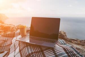 Laptop coffee on blanket with ocean view. Illustrating serene outdoor laptop use. Freelancer enjoying their time outdoors while working or browsing the internet. photo