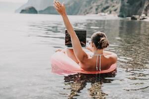 A woman is floating on a pink inflatable raft in the ocean while using a laptop. Concept of relaxation and leisure, as the woman enjoys her time in the water while working on her laptop. photo