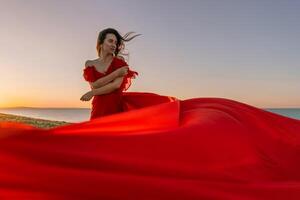 woman red dress standing grassy hillside. The sun is setting in the background, casting a warm glow over the scene. The woman is enjoying the beautiful view and the peaceful atmosphere. photo