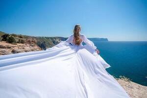 Blonde with long hair on a sunny seashore in a white flowing dress, rear view, silk fabric waving in the wind. Against the backdrop of the blue sky and mountains on the seashore. photo