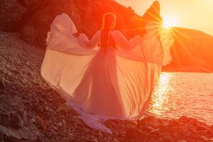 Mysterious woman silhouette long hair walks on the beach ocean water, sea nymph wind listens to the wave. Throws up a long white dress, a divine sunset. Artistic photo from the back without a face