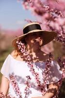 Woman blooming peach orchard. Against the backdrop of a picturesque peach orchard, a woman in a long white dress and hat enjoys a peaceful walk in the park, surrounded by the beauty of nature. photo