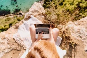 A woman is sitting on a rock overlooking the ocean with a laptop in front of her. She is enjoying the view and the peacefulness of the location. photo