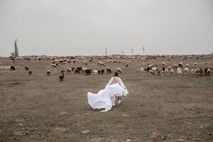 A woman in a white dress is walking through a field of cows. The cows are scattered throughout the field, with some closer to the woman and others further away. The scene has a peaceful. photo