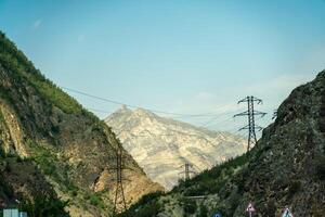 A mountain range with a power line running through it. The power line is high above the ground and is surrounded by trees. The mountains are covered in green foliage and the sky is clear and blue. photo