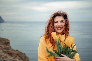 Portrait of a happy woman with hair flying in the wind against the backdrop of mountains and sea. Holding a bouquet of yellow tulips in her hands, wearing a yellow sweater photo