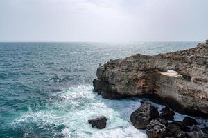The ocean is calm and the sky is cloudy. The water is a deep blue color. The rocks are jagged and the waves are small. photo