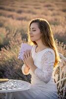 un mujer en un blanco vestir es sentado a un mesa con un libro en su regazo. ella es en un pacífico y relajado estado, posiblemente leyendo el libro o meditando el escena es conjunto en un campo de lavanda. foto