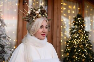Happy blonde as Snow Maiden in white costume. Wooden house exterior Festive setting. Character with crown creating festive scene. photo