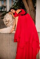 A woman in a red dress is sitting on a stone lion statue. The statue is white and has a lion's head. The woman is wearing a black belt and is looking at the camera. The scene has a calm. photo