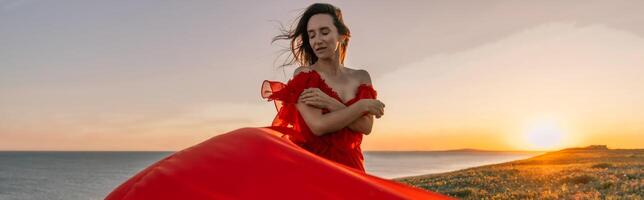 woman red dress standing grassy hillside. The sun is setting in the background, casting a warm glow over the scene. The woman is enjoying the beautiful view and the peaceful atmosphere. photo