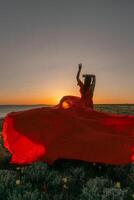 A woman in a red dress is standing in a field with the sun setting behind her. She is reaching up with her arms outstretched, as if she is trying to catch the sun. The scene is serene and peaceful. photo