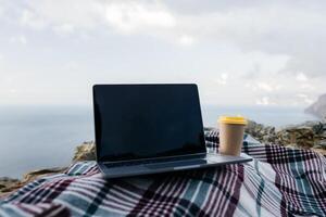 Laptop coffee on blanket with ocean view. Illustrating serene outdoor laptop use. Freelancer enjoying their time outdoors while working or browsing the internet. photo