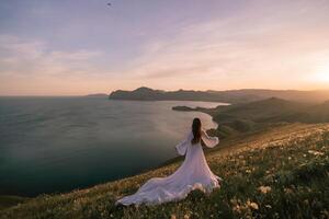 A woman in a white dress stands on a hill overlooking the ocean photo