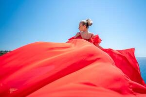 Blonde with long hair on a sunny seashore in a red flowing dress, back view, silk fabric waving in the wind. Against the backdrop of the blue sky and mountains on the seashore. photo