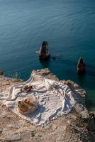 A white blanket is spread out on a beach near the water. The blanket is on a rock and there are some oranges and apples on it. The scene is peaceful and relaxing, with the ocean in the background. photo