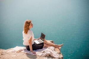 Freelance woman working on a laptop by the sea, typing away on the keyboard while enjoying the beautiful view, highlighting the idea of remote work. photo