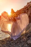 Mysterious woman silhouette long hair walks on the beach ocean water, sea nymph wind listens to the wave. Throws up a long white dress, a divine sunset. Artistic photo from the back without a face