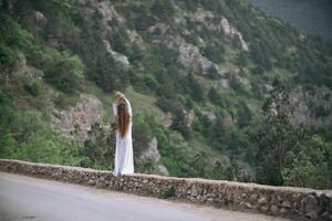 A woman stands on a stone wall overlooking a mountain. She is wearing a white dress and she is in a contemplative mood. photo