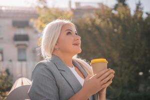 A blonde woman is sitting on a bench and holding a cup of coffee. She is smiling and looking up at the sky. photo