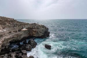 A rocky cliff overlooks the ocean, with the water crashing against the rocks. The scene is serene and peaceful, with the sound of the waves providing a calming atmosphere. photo