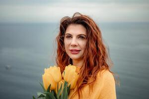 Portrait of a happy woman with hair flying in the wind against the backdrop of mountains and sea. Holding a bouquet of yellow tulips in her hands, wearing a yellow sweater photo