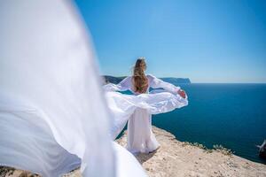 Blonde with long hair on a sunny seashore in a white flowing dress, rear view, silk fabric waving in the wind. Against the backdrop of the blue sky and mountains on the seashore. photo