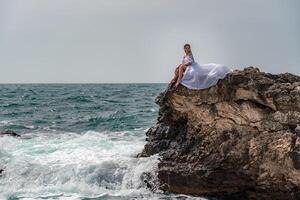 A woman in a storm sits on a stone in the sea. Dressed in a white long dress, waves crash against the rocks and white spray rises. photo