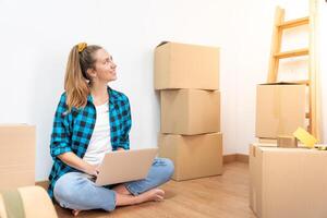 Young happy woman sitting on the floor with many boxes, working with computer in a new house. photo