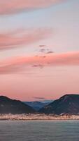 Pink clouds in evening sky over blue sea on background of city and mountains on horizon, vertical frame. photo