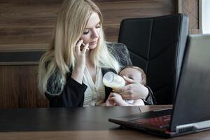 Young business woman feeds baby from bottle in office and talks on phone, concept of combining business and caring for newborn baby. photo