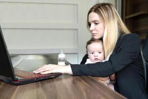 Young woman holds baby in her arms, in office, combining this with working on laptop., concept of combining business and caring for newborn baby photo