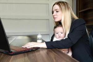 Concept of combining business and caring for newborn baby. young businesswoman holds baby in her arms, in office, combining this with working on laptop. photo
