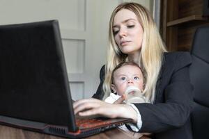 Concept of combining business and caring for a newborn child, young business woman feeds a baby from bottle in office, combining it with work on laptop. photo