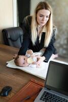 Young business woman feeds baby from bottle on desktop in office, combining it with work on laptop, vertical frame photo