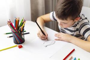 A child boy draws on white paper with colored pencils while sitting at a table photo