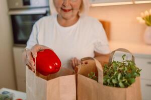 Happy Caucasian 60s woman sort vegetables from eco package in minimalist kitchen. Online buying food and grocery. Delivery service and mobile app. photo