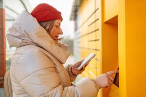 Smiling beautiful Caucasian woman client holding cardboard boxes and hands show smartphone using automated self service post terminal machine or locker. Mail shipping concept. photo