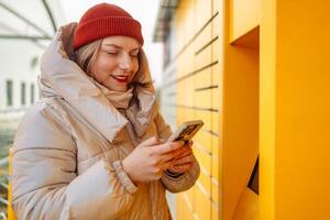 Happy smiling woman client with box at outdoor automated parcel machine choosing operation on touch screen by smart phone photo