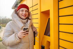 Happy smiling woman client with box at outdoor automated parcel machine choosing operation on touch screen by smart phone photo