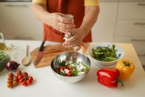 Close-up of a 60s woman preparing healthy vegetable salad in kitchen, seasoning meal, adding some herbs and salt to bowl, enjoying cooking vegetarian food at home, free space for advertising text photo