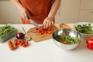 Hands of senior 60s female chopping fresh tomato on wooden board while preparing italian salad with vegetables photo