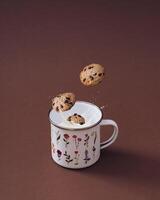 Chocolate chip cookies falling into vintage floral enamel mug full of fresh milk on brown background. Sweet food biscuit concept. Minimal layout. Trendy chocolate cookies with milk composition. photo