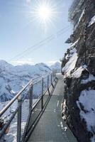 Metal walkway on rocky cliff at Murren ski resort, Switzerland. Snow covered mountains in distance, clear blue sky, and safe pathway for exploring Alps. photo