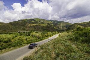 Car driving on country road with mountains, grass, and clouds in the background photo