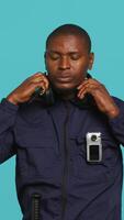 Vertical Security guard equipped with bodycam listening music, taking break from job shift. Portrait of bodyguard walking around perimeter with headphones on while at work, studio background, camera A video