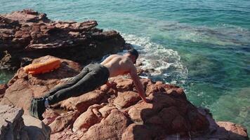 A man does push-ups from the floor on rocks by the sea video
