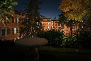 Nighttime scene at a luxury restaurant. Empty outdoor table surrounded by lush vegetation. Grand building facade in background. Serene, upscale ambiance. photo