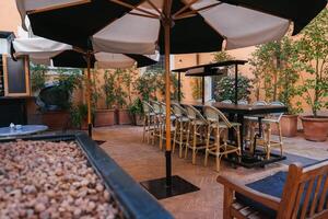 Luxury hotel outdoor seating area in Rome. Warm ambiance with terracotta tiles, cream umbrellas, rattan chairs, lush plants. Elegant and tranquil setting. photo