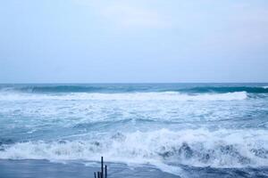 As the tide sweeps in at South Jogja Jetis Beach, Cornwall under a serene sky, waves gently roll, accompanied by ripples and bubbling foam. photo